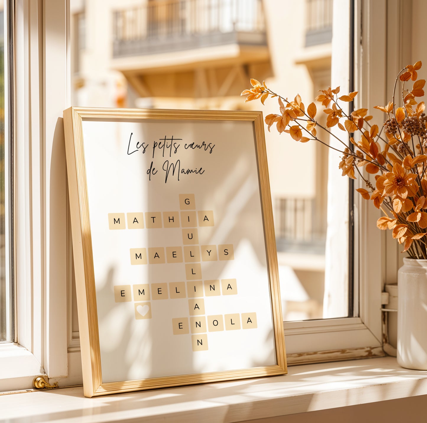 Affiche Mamie Personnalisée in gold frame with Scrabble-style names for Fête des Grands-Mères, displayed by window with.