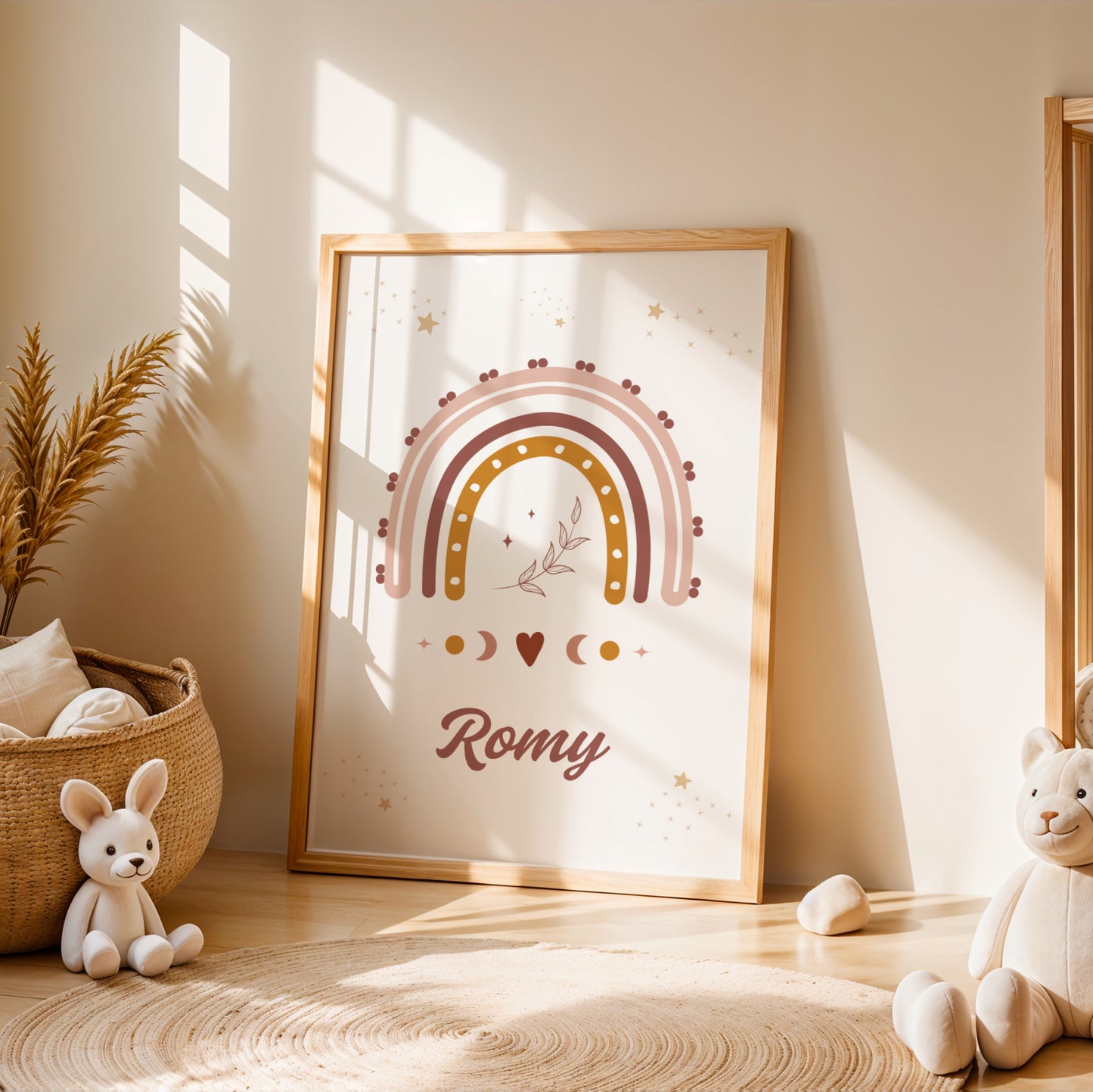 Une affiche personnalisée pour chambre d'enfant posée au sol dans un cadre en bois clair, entourée de peluches. L'illustration représente un grand arc-en-ciel bohème aux couleurs terracotta, rose et ocre, surmonté de petites étoiles dorées et d'un brin de feuillage délicat en son centre. Sous l'arc-en-ciel, des symboles de phases lunaires et un petit cœur entourent le prénom "Romy" écrit dans une typographie élégante et arrondie de couleur chocolat.