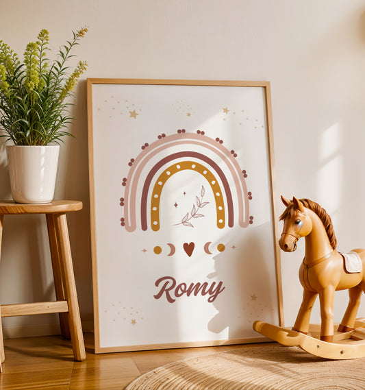 Une affiche personnalisée pour chambre d'enfant posée au sol dans un cadre en bois clair, entourée de peluches. L'illustration représente un grand arc-en-ciel bohème aux couleurs terracotta, rose et ocre, surmonté de petites étoiles dorées et d'un brin de feuillage délicat en son centre. Sous l'arc-en-ciel, des symboles de phases lunaires et un petit cœur entourent le prénom "Romy" écrit dans une typographie élégante et arrondie de couleur chocolat.