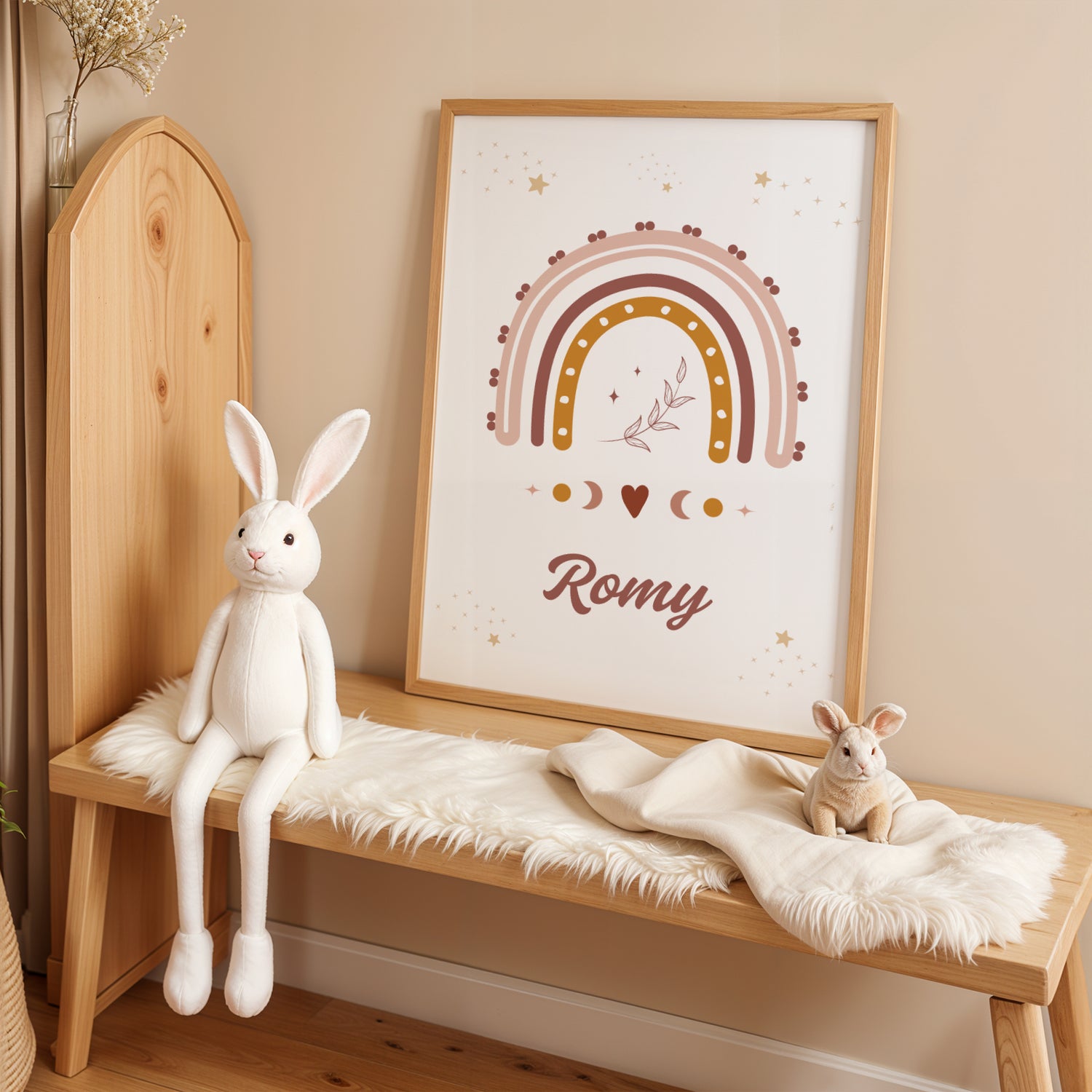 Une affiche personnalisée pour chambre d'enfant posée au sol dans un cadre en bois clair, entourée de peluches. L'illustration représente un grand arc-en-ciel bohème aux couleurs terracotta, rose et ocre, surmonté de petites étoiles dorées et d'un brin de feuillage délicat en son centre. Sous l'arc-en-ciel, des symboles de phases lunaires et un petit cœur entourent le prénom "Romy" écrit dans une typographie élégante et arrondie de couleur chocolat.
