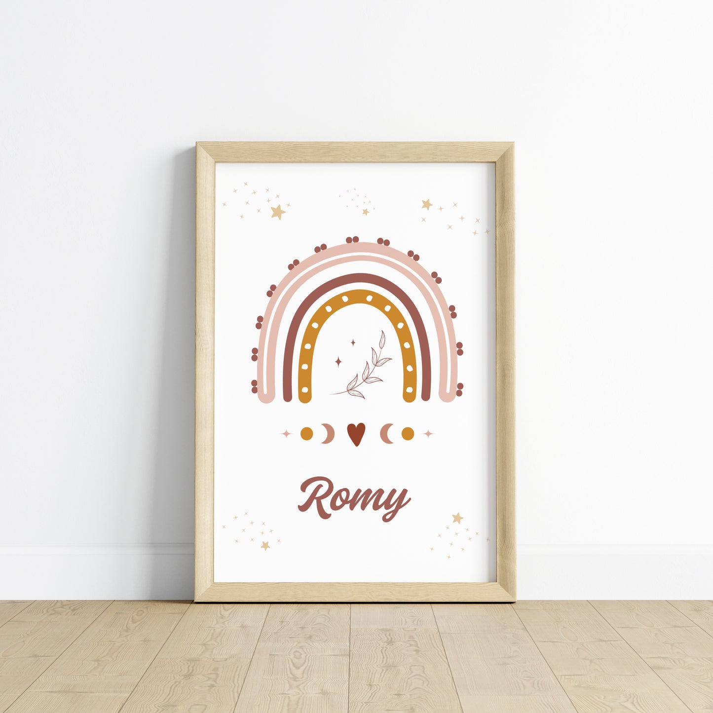 Une affiche personnalisée pour chambre d'enfant posée au sol dans un cadre en bois clair, entourée de peluches. L'illustration représente un grand arc-en-ciel bohème aux couleurs terracotta, rose et ocre, surmonté de petites étoiles dorées et d'un brin de feuillage délicat en son centre. Sous l'arc-en-ciel, des symboles de phases lunaires et un petit cœur entourent le prénom "Romy" écrit dans une typographie élégante et arrondie de couleur chocolat.