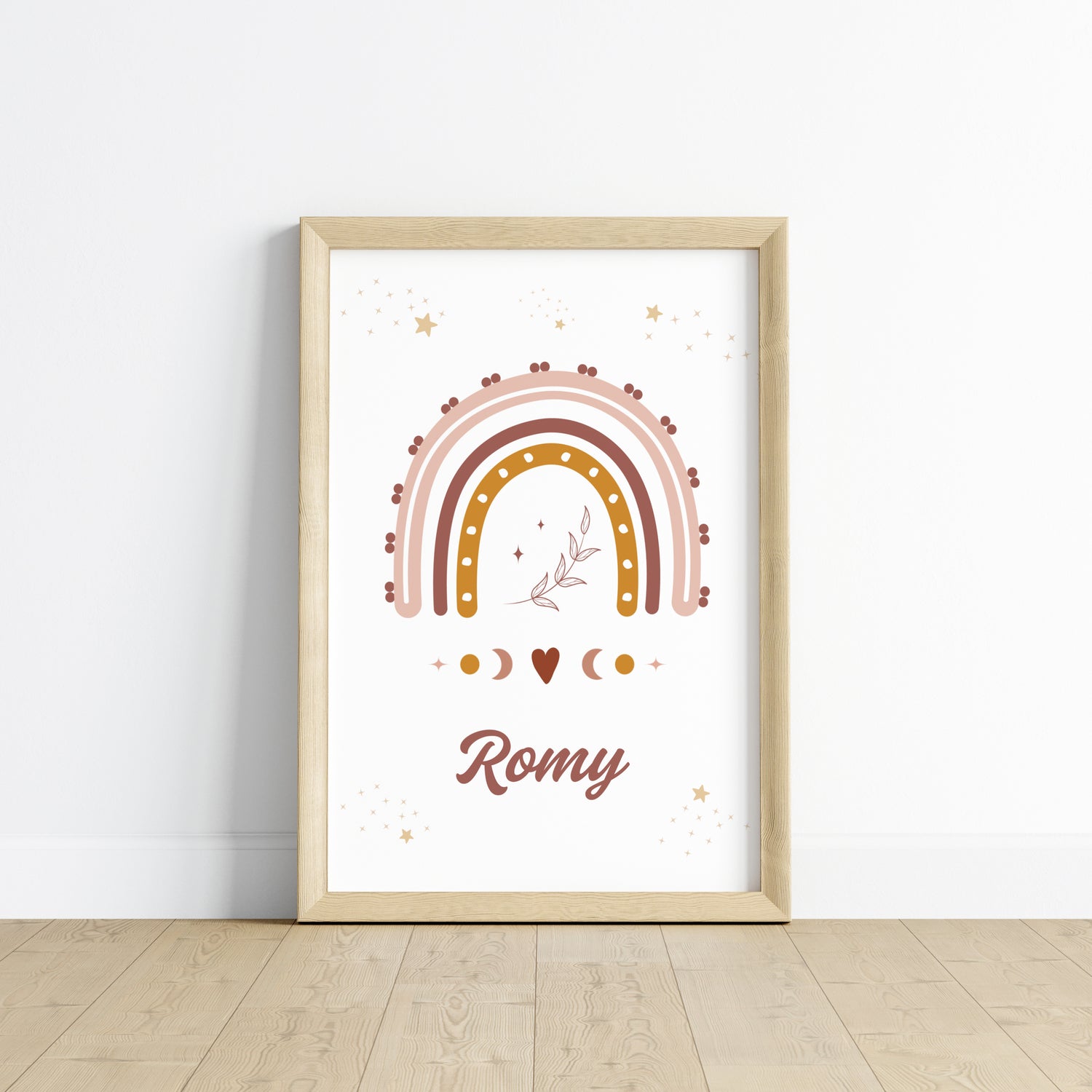 Une affiche personnalisée pour chambre d'enfant posée au sol dans un cadre en bois clair, entourée de peluches. L'illustration représente un grand arc-en-ciel bohème aux couleurs terracotta, rose et ocre, surmonté de petites étoiles dorées et d'un brin de feuillage délicat en son centre. Sous l'arc-en-ciel, des symboles de phases lunaires et un petit cœur entourent le prénom "Romy" écrit dans une typographie élégante et arrondie de couleur chocolat.